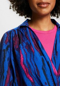 Tissu bleu texturé avec des rayures ondulées roses et noires, doté d'un col et d'un design superposé sur une chemise rose côtelée.