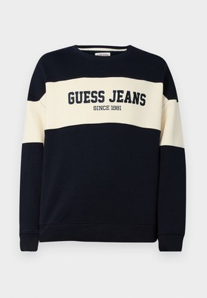 Laivastonhuppari, jossa on vaaleanruskea vaakaraita, sisältää "GUESS JEANS" -tekstin ja "SINCE 1981" -yksityiskohdan. Valmistettu pehmeästä puuvillamateriaalista.