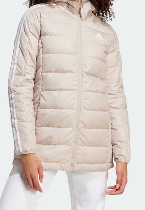 Personne portant une veste matelassée beige Adidas avec capuche et détails blancs à trois bandes sur les manches, associée à un pantalon blanc.