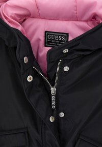 Schwarze Jacke mit pinkem Innenfutter, ausgestattet mit Reißverschluss, Druckknöpfen und einem Etikett mit der Marke Guess. Glatte äußere Textur.