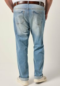 Jeans de mezclilla azul claro con pierna ajustada, zonas desgastadas y dos bolsillos traseros. Los puños enrollados revelan un forro de tela clara. Cinturón marrón como acento.