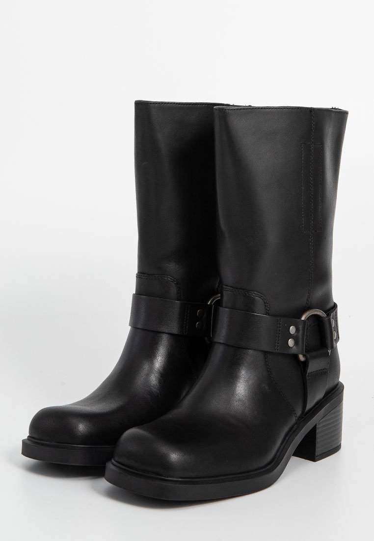 Paar schwarze Lederstiefel bis zur Mitte der Waden mit Riemen und metalischem Ringdetail, abgerundete Zehen und Blockabsätzen auf einem weißen Hintergrund.