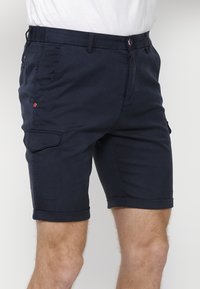 Shorts cargo bleu marine en tissu léger, avec poches latérales, ourlets roulés et fermeture à bouton avec des accents contrastants.