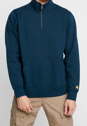 Uomo con felpa blu navy con zip a quarti e tasca frontale, indossa pantaloni cargo color sabbia, in piedi su sfondo neutro.