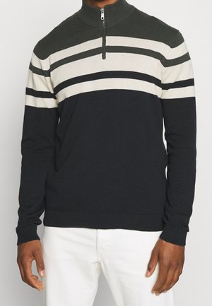Mann trägt einen schwarz, weiß und dunkelolivgrün gestreiften Halbzip-Pullover mit weißen Hosen, steht vor einem einfarbigen Hintergrund.