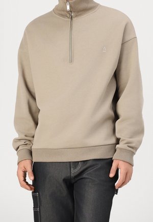 Mand iført beige sweatshirt med kvart-lynlås, ribstrikkede manchetter og kant, kombineret med sorte bukser med hvide sømme og detaljerede sidelommer.