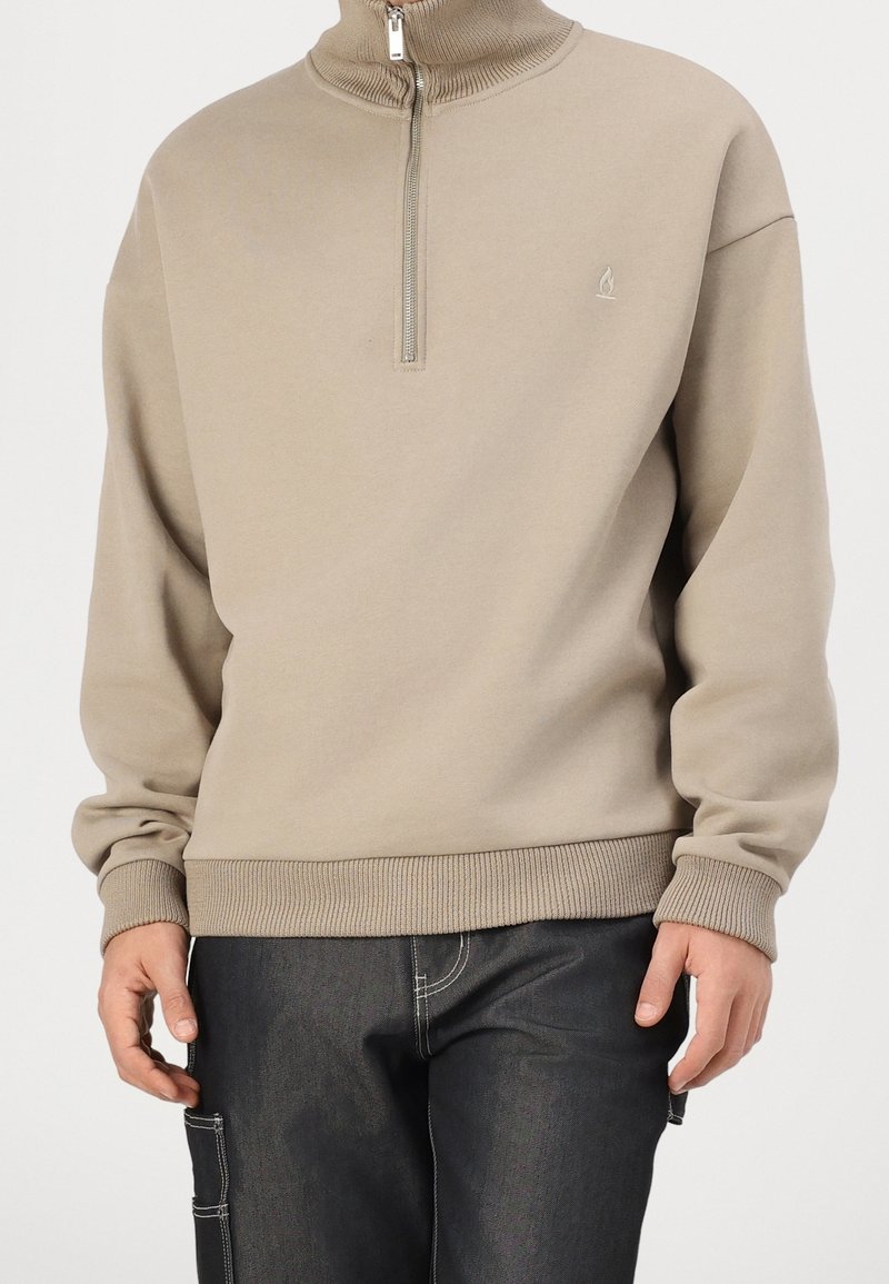 Homme portant un sweat-shirt beige à fermeture quart de zip avec poignets et ourlet côtelés, associé à un pantalon noir avec des coutures blanches et des détails de poche latérale.