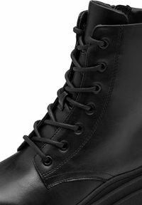 Bottine en cuir noir à bout rond, avec des coutures verticales, six œillets en métal et lacets noirs. Fini texturé sur le matériau.