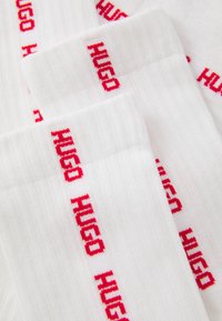 Hvide ribbede sokker med rød "HUGO" tekst gentaget lodret langs kanten, arrangeret overlappende på en flad overflade.