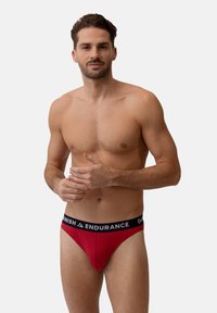Tanga rosso in cotone con vita elastica nera recante il logo "DANISH ENDURANCE". Texture liscia, forma sagomata, copertura minima.