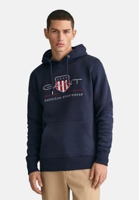 Felpa con cappuccio blu navy realizzata in misto cotone. Presenta una tasca frontale, polsini a coste e un logo grafico con un design a scudo.