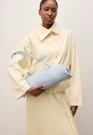 Naine, kes kannab kreemjat, suuruses nööbitavat särk-kleiti, hoides helesinist ristkülikukujulist käekotti kahekordsete käepidemetega.