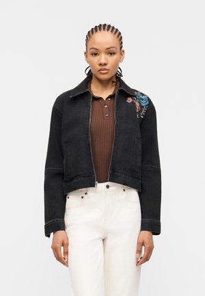 Νεαρό άτομο με πλεξούδες φοράει μαύρο cropped σακάκι με κεντήματα λουλουδιών στον ώμο, καφέ πλεκτό πόλο και λευκά παντελόνια.