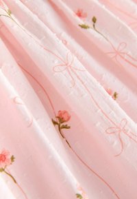 Rozā audums ar teksturētu rakstu, kura centrā ir ziedu izšuvumi rozā un zaļā krāsā, kā arī maigas bantiņas, ieskaitītas pasteļa rozā diegos.