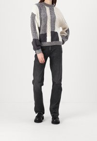 Pull en tricot avec un mélange de blocs de couleurs gris et crème, motifs texturés, et un col rond, associé à un jean foncé et des chaussures noires.