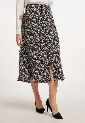 Vrouw draagt een zwarte midirok met bloemenprint en een vooraan gemaakte split, een witte blouse en zwarte hoge hakken, staand tegen een effen achtergrond.
