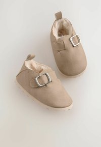 Tan ruskind babysko med en blød foring, der har en spænde i siden og en struktureret gummisål. Rund tå og med en strop bagpå.