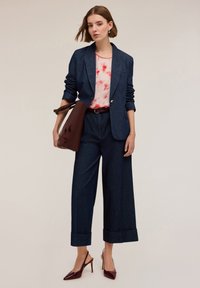 Dżinsowa marynarka nałożona na różowo-czerwony top w stylu tie-dye, połączona z szerokimi dzinsowymi spodniami w ciemnym kolorze oraz burgundowymi sandałkami na obcasie. Brązowa torebka jako dodatek.