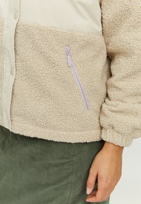 Beige fleece jacket with a cream upper section, featuring a side zip pocket with a lavender zipper and elasticated cuffs.