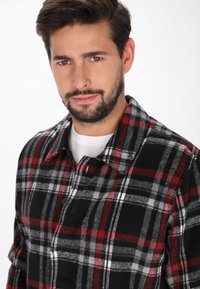 Svart och röd rutig flanellskjorta med knäppning, utrustad med krage och en mjuk textur. Modellen visar skjortan mot en vit bakgrund.