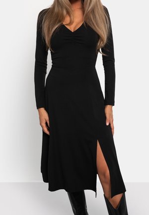 Frau trägt ein langärmliges schwarzes Kleid mit einem Seitenschlitz, kombiniert mit schwarzen kniehohen Stiefeln, und steht vor einem schlichten weißen Hintergrund.