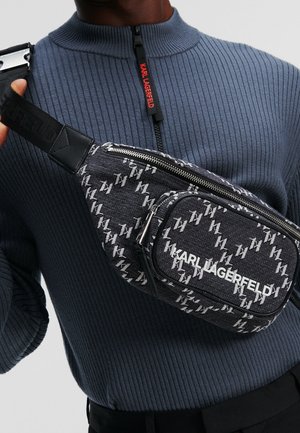 Mann trägt einen blau gerippten Reißverschluss-Pullover und eine schwarze Umhängetasche mit weißem Karl Lagerfeld Monogramm-Muster und Logo.