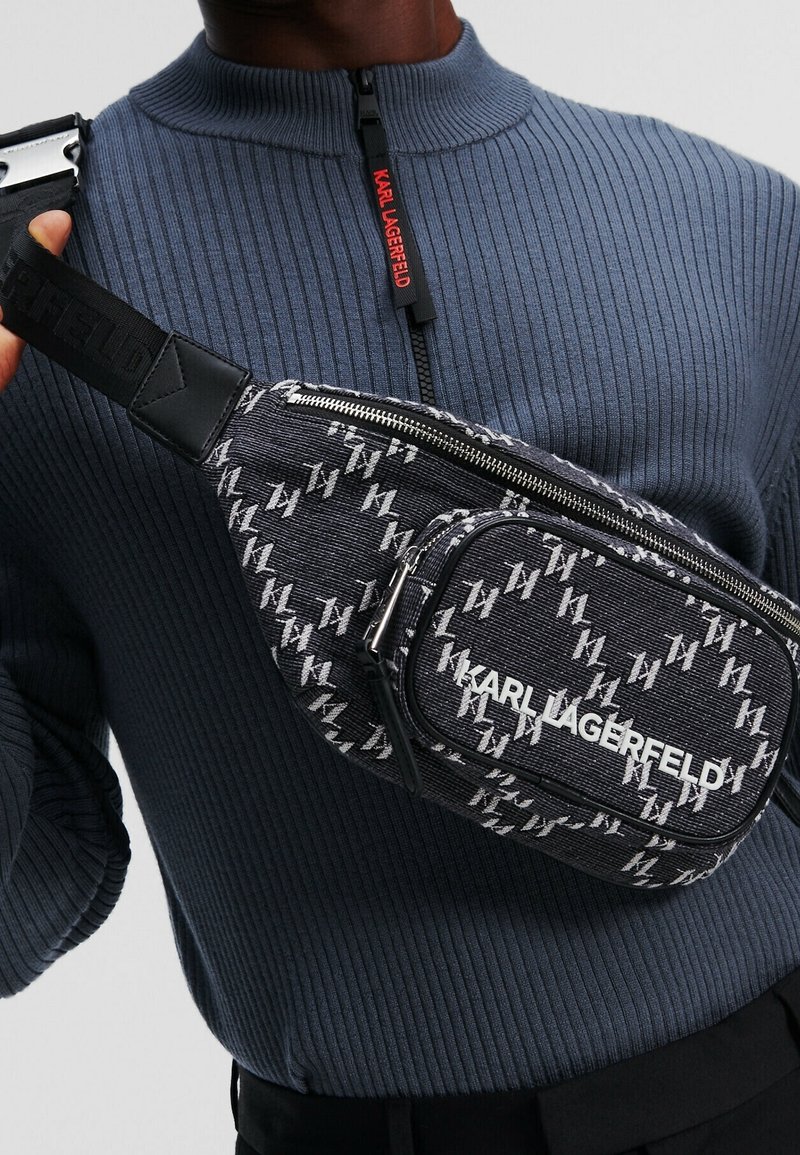 Homme portant un pull bleu zippé côtelé et un sac bandoulière noir avec un motif monogramme blanc Karl Lagerfeld et un logo.