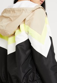 Chaqueta con bloques de color que presenta paneles en beige, blanco, amarillo claro y negro. Confeccionada en material brillante, cuenta con capucha y cintura elástica.