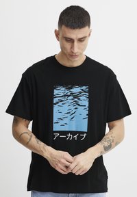 Juodas medvilninis marškinėlis su mėlynų vandens bangų grafika ir japonišku tekstu. Trumpomis rankovėmis ir laisvo silueto dizainas.