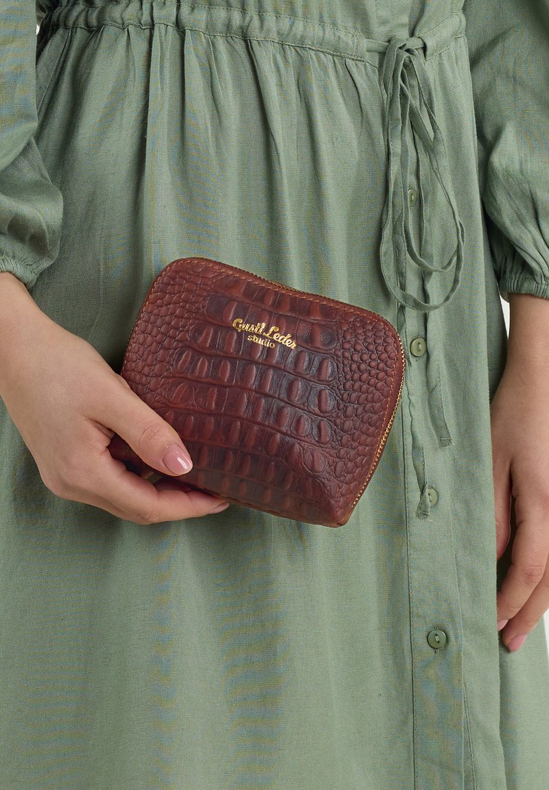 Hnedá kožená peňaženka s textúrovaným krokodílím vzorom a zlatým logom. Kompaktný dizajn držiaci sa v jednej ruke proti zelenej šaty.