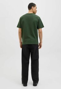 Grøn kortærmet t-shirt med en struktureret overflade, parret med sorte, løstsiddende bukser. Baggrunden er ensfarvet hvid.