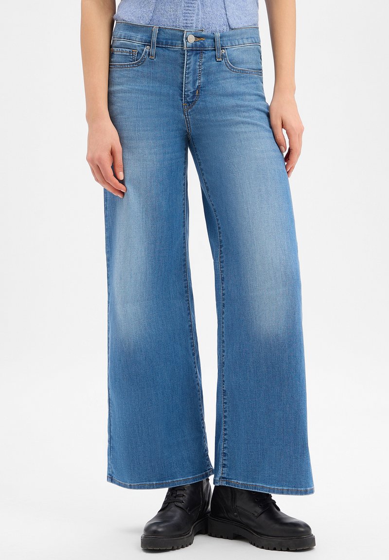 Light blue wide-leg jeans worn with black lace-up boots, shown from waist to feet on a person with hands relaxed by sides.