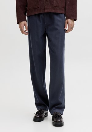 Άνδρας που φοράει σκούρο ναυτικό μπλε παντελόνι με ίσια γραμμή, σκουρόκαφέ μοκασίνια με φούντες και μπουφάν από μαρόν κολάρο ύφασμα κορδούρας.