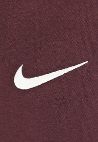 Logo swoosh Nike ricamato bianco su sfondo di tessuto testurizzato borgogna.