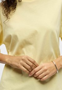 Personne portant une chemise jaune pâle, mains doucement jointes, avec un bracelet argenté au poignet droit et des cheveux bouclés visibles sur le côté gauche.