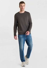 Pull en maille gris foncé, col rond, manches longues ; associé à un jean en denim bleu clair et des baskets beiges. Le mannequin se tient sur un fond neutre.