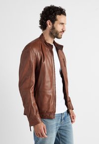 Braune Lederjacke mit Stehkragen, Frontreißverschluss, seitlichen Taschen und genähten Details, getragen über einem weißen Hemd und blauen Jeans.