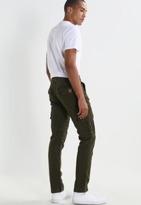 Pantalones cargo de color verde oliva con múltiples bolsillos, piernas ajustadas y un parche de tela en la parte trasera; combinados con una camiseta blanca ajustada y zapatillas blancas.