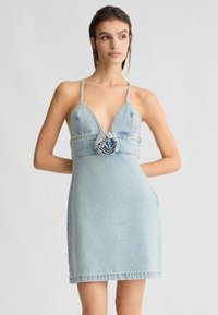 Abito in denim azzurro chiaro, con scollatura a V profonda, spalline sottili e un'applicazione stilizzata a forma di fiore in vita. Silhouette ad A, lunghezza corta.