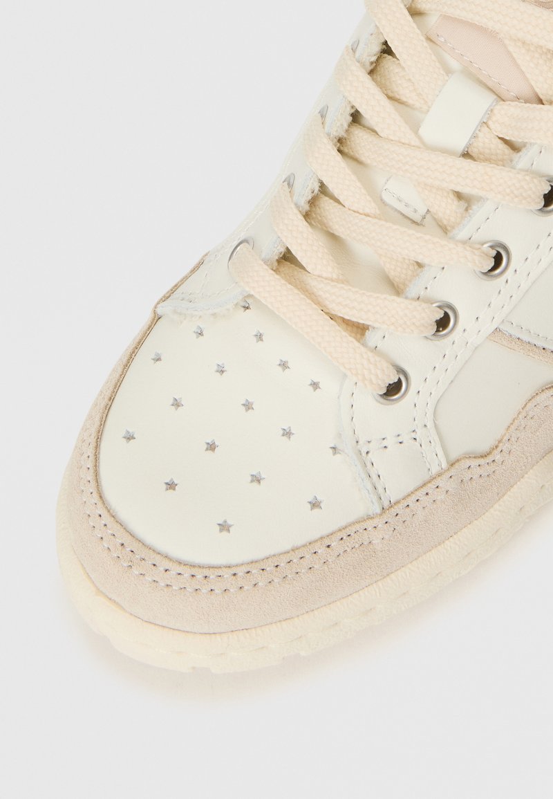Primer plano de la puntera de una zapatilla beige con perforaciones en forma de estrella, ribete de ante y cordones crema pasados por ojales de metal.