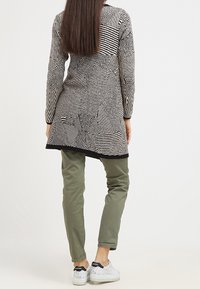 Svart- och vitmönstrad lång topp med geometriska mönster, kombinerad med olivgröna byxor och vita sneakers, med svarta accenter.