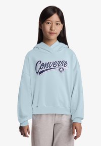 Φόρμα με κουκούλα σε ανοιχτό γαλάζιο, με χαλαρή εφαρμογή, που διαθέτει εμφανές λογότυπο "Converse" και το κλασικό αστέρι. Το υλικό φαίνεται μαλακό και υφασμάτινο.