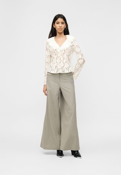 Γυναίκα που φοράει λευκό μπλουζάκι από δαντέλα με λουλούδια και γιακά με βολάν, μπεζ παντελόνι φαρδιάς γραμμής και μαύρες μπότες με τακούνι σε απλό φόντο.