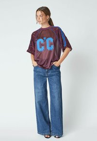 Maroon mesh top with lace sleeves, featuring blue graphic "C'C" design. Paired with wide-leg blue denim jeans and black heeled sandals.