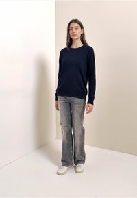 Marineblauer Strickpullover mit Rundhalsausschnitt, kombiniert mit hellgrauen ausgestellten Jeans und weißen Turnschuhen. Glatte Textur, lässiges Design.