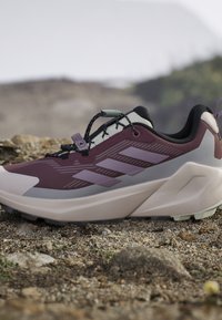 Trailrunning-Schuh in Lila und Grau mit gedämpfter Sohle und schnellem Schnürsystem auf felsigem Outdoor-Gelände.