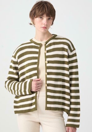 Νεαρός με κοντά μαλλιά φοράει ένα ζακέτα με ρίγες σε ελιά πράσινο και λευκό με κουμπιά πάνω από μια μπεζ μπλούζα και παντελόνι, σε απλό φόντο.