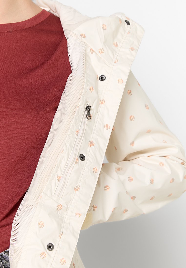 Lichte, crème-kleurige jas met een open gaasvoering en oranje spiraalpatronen. Voorzien van een ritsluiting en drukknoopjes.