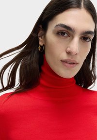 Maglione a collo alto rosso con un design aderente e texture a coste. Orecchini a cerchio oro che accentuano il look. Tessuto naturale e morbido.