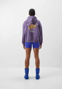 Fioletowa bluza z kapturem z żółtą kwiatową biżuterią i napisami „równość” oraz „jedność”. Do tego niebieskie spodenki i niebieskie buty.